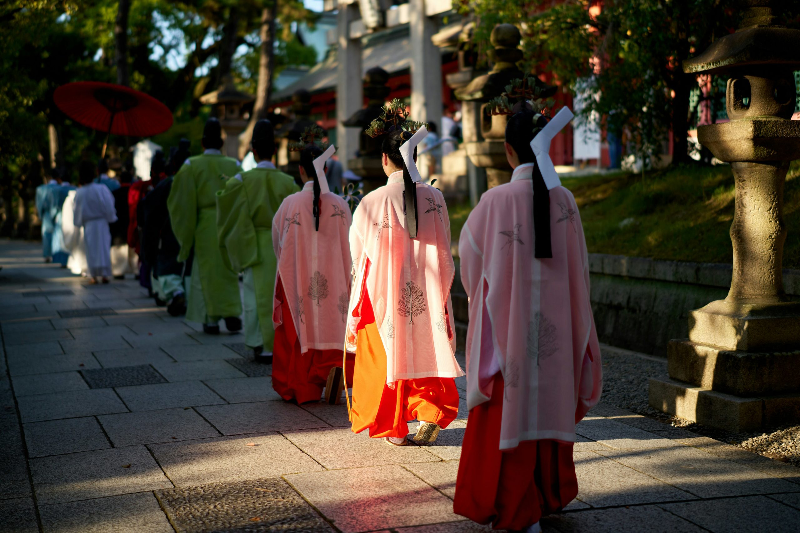Traditional Japanese ceremonial procession with people in vibrant pink and orange kimono attire walking through spiritual setting with stone lanterns and greenery