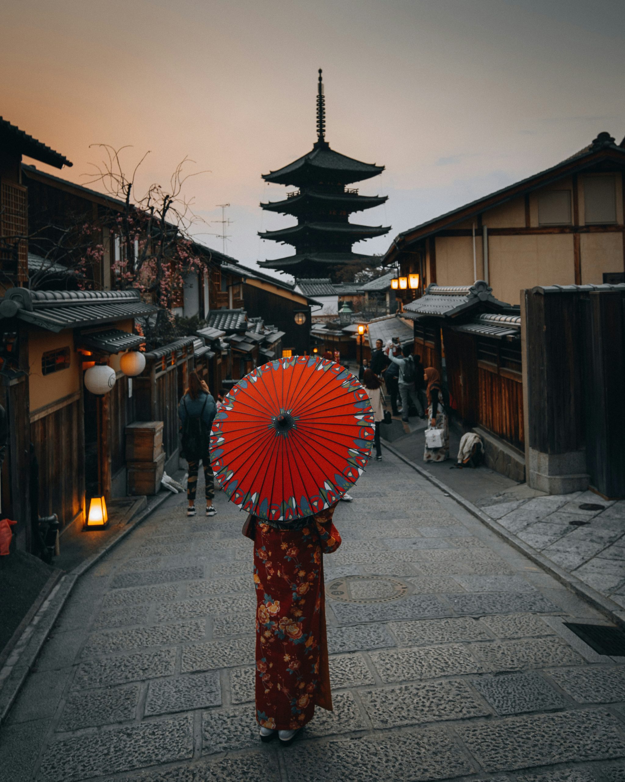 Traditional kimono-clad woman with red parasol walking through historic Kyoto street with pagoda in background at dusk showcasing the integration of traditional dress with sacred spaces