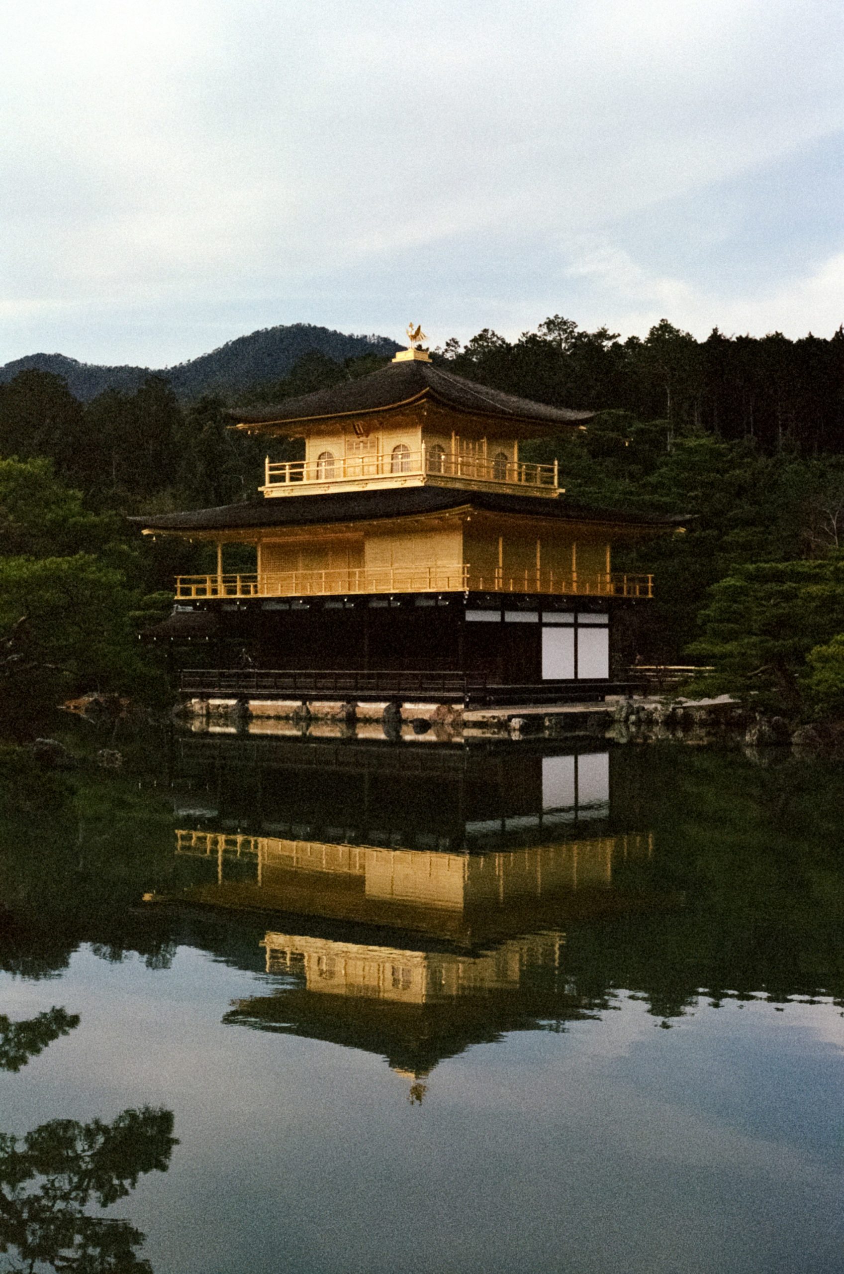Kinkaku-ji Golden Pavilion temple in Kyoto reflected in serene pond waters, surrounded by lush green trees during golden hour, representing the timeless beauty of traditional Japanese architecture and cultural heritage