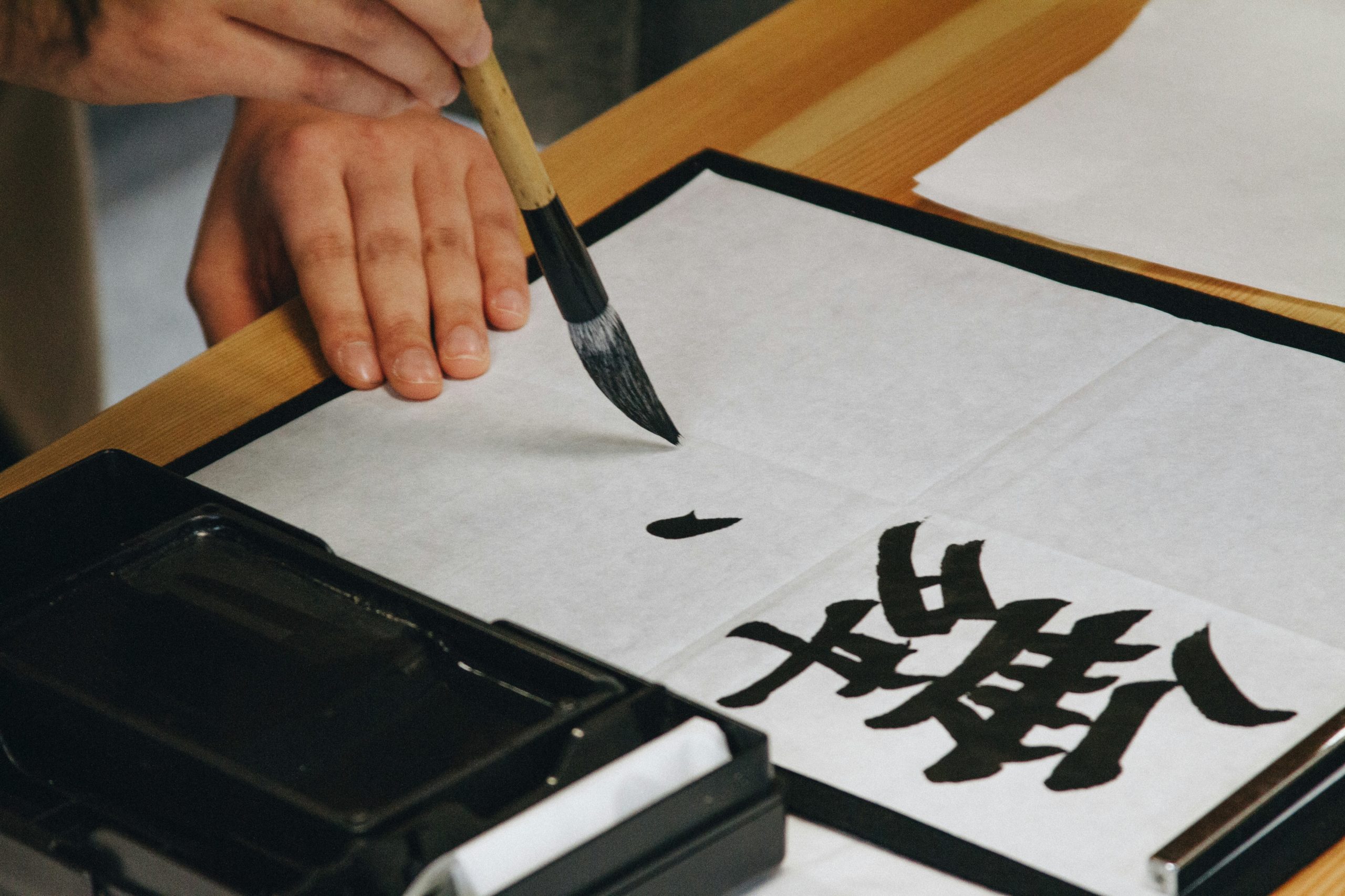 Close-up view of traditional Japanese calligraphy (shodō) being written with a black ink brush on white paper, demonstrating the elegant brushstrokes and artistic precision of Japanese writing